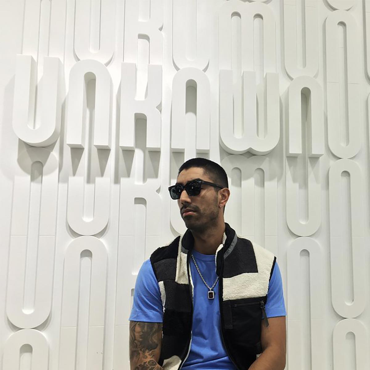 Man wearing sunglasses inside, in front of a wall with Unknwn in relief.
